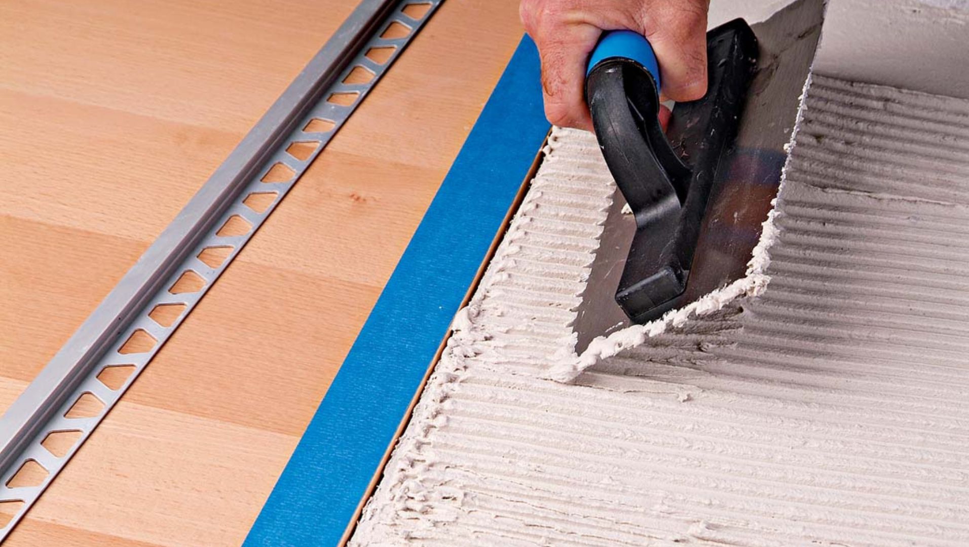 A worker installing tile trim on a tiled wall, using tools and adhesive.